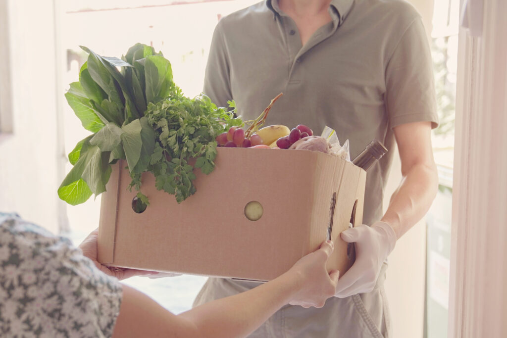 A person holding a box full of fresh produce and canned goods hands it to another person out of frame.